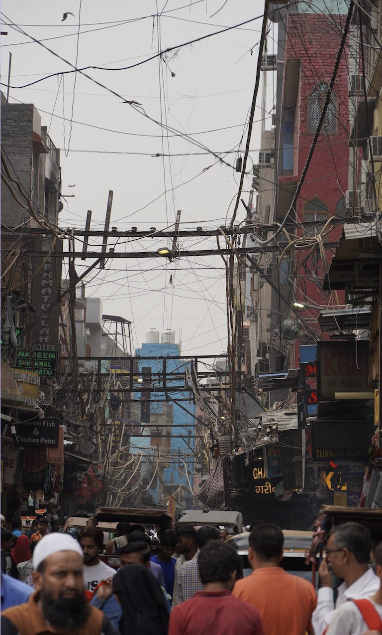 A photograph of Chandni Chowk in Delhi, India, where blurred crowds move below tangled overhead wires. While people pass by disconnected, these wires symbolise the invisible digital threads that connect us—suggesting how modern connection often exists beyond physical presence.